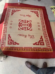 Hình ảnh chụp một tấm chiếu cói được trải trên nền xi măng. Chiếu có màu be tự nhiên, viền và họa tiết trang trí màu đỏ. Trên chiếu có in chữ nghệ thuật màu đỏ: 'Nga Sơn' và 'Kỷ Niệm'. Chính giữa chiếu là một biểu tượng hình tròn lớn màu đỏ, được thiết kế theo kiểu chữ triện cách điệu. Xung quanh có thể thấy chân và tay của một vài người, cho thấy họ đang xem hoặc trưng bày tấm chiếu. Đây là một sản phẩm thủ công truyền thống đặc trưng của vùng Nga Sơn.。（日本語訳）。セメントの床の上に敷かれた、い草のござを写した写真。ござは自然なベージュ色で、縁と装飾模様は赤色です。ござの上には、「Nga Sơn」と「Kỷ Niệm」という赤い芸術的な文字が印刷されています。ござの中央には、様式化された篆書体風にデザインされた、大きな円形の赤いシンボルがあります。周りには数人の足や手が見え、彼らがそのござを見ているか、または展示していることを示しています。これはガソン地方特有の伝統的な手工芸品です。