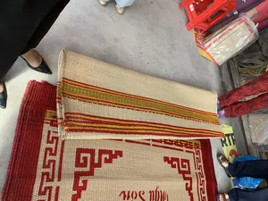 Bối cảnh là một cửa hàng hoặc xưởng sản xuất, nơi mọi người đang xem và lựa chọn chiếu cói. Trên sàn nhà xi măng, một chiếc chiếu cói được trải ra một phần, có viền và hoa văn hình học màu đỏ. Trên chiếu có dệt dòng chữ "Nga Sơn". Một chiếc chiếu khác được cuộn lại, có hoa văn sọc nhiều màu (đỏ, vàng, xanh lá), đặt trên chiếc chiếu trải. Xung quanh có chân của nhiều người đi các loại giày dép khác nhau, cho thấy họ đang tụ tập để mua sắm. Ở phía sau có một chiếc ghế nhựa màu đỏ và những chiếc chiếu khác được đóng gói trong túi ni lông.。（日本語訳）。背景は店か工房で、人々がい草のござを見て選んでいます。セメントの床の上には、一枚のい草のござが部分的に広げられており、赤い縁取りと幾何学模様があります。ござには「Nga Sơn」という文字が織り込まれています。別のござは巻かれており、多色（赤、黄、緑）の縞模様があり、広げられたござの上に置かれています。周りには様々な履物を履いた多くの人々の足があり、彼らが買い物をするために集まっていることを示しています。後ろには赤いプラスチックの椅子と、ビニール袋に梱包された他のござがあります。