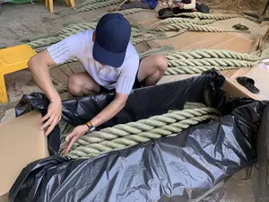 Trong ảnh, một người đàn ông đang làm việc tại một cơ sở sản xuất thủ công, có thể là một xưởng gia đình. Anh đang ngồi xổm và cẩn thận xếp một cuộn dây thừng rất lớn, được bện từ sợi thực vật tự nhiên (giống như sợi cói), vào một thùng carton đã được lót bằng túi ni lông đen. Người đàn ông mặc áo phông trắng, quần short và đội một chiếc mũ lưỡi trai màu xanh đậm có thêu chữ 'NATIONAL GEOGRAPHIC'. Trên sàn bê tông xung quanh, có nhiều cuộn dây thừng thành phẩm khác được đặt trên những tấm chiếu cói. Ở hậu cảnh, có những người khác cũng đang ngồi làm công việc tương tự. Một chiếc ghế nhựa màu vàng được đặt ở phía bên trái.。（日本語訳）。写真には、一人の男性が手工業の生産施設で働いています。それは家内工場かもしれません。彼はしゃがみ込み、天然の植物繊維（イグサの繊維のようなもの）で編まれた非常に大きな縄の巻物を、黒いビニール袋が敷かれた段ボール箱に慎重に詰めています。その男性は白いTシャツと短パンを着用し、「NATIONAL GEOGRAPHIC」という文字が刺繍された濃い青色の野球帽をかぶっています。周りのコンクリートの床の上には、イグサのござの上に置かれた、完成品の縄の巻物が他にもたくさんあります。背景には、他の人たちも座って同様の作業をしています。左側には黄色いプラスチックの椅子が一つ置かれています。