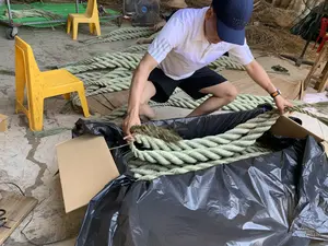 Hình ảnh ghi lại cảnh một người đàn ông đang làm việc trong một không gian trông giống như một xưởng sản xuất thủ công. Người đàn ông mặc áo phông polo trắng có logo "adidas", quần đùi đen và đội mũ lưỡi trai màu xanh đậm, đang ngồi xổm trên sàn nhà. Anh ấy đang sắp xếp những cuộn dây thừng lớn, được bện từ sợi cói (hoặc một loại thực vật tương tự), vào một chiếc thùng carton lớn có lót túi ni lông màu đen. Xung quanh có nhiều vật dụng liên quan đến công việc: những chiếc ghế nhựa màu vàng, các bó sợi cói khô nằm trên sàn và trên một chiếc xe đẩy có lưới màu xanh lá cây ở phía sau. Ngoài ra còn có một chiếc quạt điện và một người khác đội nón đang ngồi làm việc ở hậu cảnh. Toàn bộ khung cảnh cho thấy đây là một công đoạn trong quy trình sản xuất hoặc đóng gói các sản phẩm thủ công từ cói.。（日本語訳）。写真は、手工芸の作業場のような空間で一人の男性が作業している光景を捉えている。その男性は、「adidas」のロゴが入った白いポロシャツを着て、黒い短パンを履き、濃い青色の野球帽をかぶり、床にしゃがみ込んでいる。彼は、い草（またはそれに類する植物）の繊維から編まれた大きなロープの巻物を、黒いビニール袋が敷かれた大きな段ボール箱に詰めている。周囲には仕事に関連する多くの物がある：黄色いプラスチックの椅子、床や後方にある緑色の網が付いた台車の上に置かれた乾燥したい草の繊維の束。さらに、扇風機と、背景には笠をかぶって座って作業している別の人物もいる。全体の光景は、これがい草から作られた手工芸品の製造または梱包プロセスにおける一工程であることを示している。