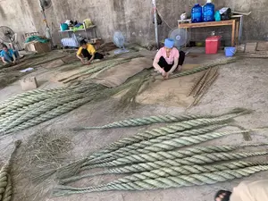 Bối cảnh là một xưởng sản xuất thủ công với sàn bê tông. Có ba người phụ nữ đang ngồi trên sàn nhà, trên những tấm chiếu, để làm việc. Họ đang thực hiện công việc bện các sợi thực vật khô (giống như sợi cói) để tạo thành những sợi dây thừng rất lớn và dày. Rất nhiều cuộn dây thừng đã hoàn thành được xếp trên sàn. Xung quanh nơi làm việc có các vật dụng như quạt điện, một chiếc bàn gỗ đựng các bình nước uống lớn màu xanh, một thùng đá màu đỏ và một chiếc xô nhựa màu xanh. Không có chữ viết nào có thể đọc được trong hình ảnh.。（日本語訳）。背景はコンクリートの床の手工業の作業場です。3人の女性が、作業をするために、床のござの上に座っています。彼女たちは、乾燥した植物繊維（い草の繊維のようなもの）を編んで、非常に大きくて太いロープを作る作業をしています。完成したロープの巻物がたくさん床に積まれています。作業場の周りには、扇風機、青い大きな飲料水ボトルが置かれた木製のテーブル、赤いクーラーボックス、そして青いプラスチックのバケツのような物があります。画像の中には、読み取れる文字はありません。