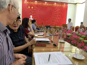 Bản ghi lại thông tin văn bản: Trên tấm phông nền màu đỏ có dòng chữ lớn màu vàng: 'ĐẢNG CỘNG SẢN VIỆT NAM QUANG VINH MUÔN NĂM。'. Trên tài liệu đặt trên bàn có thể đọc được các dòng chữ: 'CỘNG HÒA XÃ HỘI CHỦ NGHĨA VIỆT NAM, Độc lập - Tự do - Hạnh phúc', 'Nga Sơn, ngày 13 tháng 6 năm 2023', 'Kính gửi: Sở Công Thương tỉnh Thanh Hóa', và nội dung là báo cáo của UBND huyện Nga Sơn về tình hình phát triển làng nghề.。Mô tả bối cảnh và hoạt động: Bức ảnh chụp cảnh một cuộc họp đang diễn ra trong một phòng họp trang trọng. Nhiều người, bao gồm cả những người lớn tuổi, đang ngồi quanh một chiếc bàn họp dài, bóng loáng. Một người đàn ông mặc áo sơ mi trắng đang đứng phát biểu trước mọi người. Những người tham dự khác đang tập trung lắng nghe. Trên bàn có đặt sẵn tài liệu, sổ tay, máy tính xách tay, micro, chai nước và cốc thủy tinh. Ở giữa bàn được trang trí bằng một dãy hoa sen hồng. Phía sau người phát biểu là một tấm phông nền lớn màu đỏ, trên đó có hình trống đồng Đông Sơn, biểu tượng Đảng Cộng sản Việt Nam (búa liềm và ngôi sao), và một bức tượng bán thân của Chủ tịch Hồ Chí Minh đặt ở phía trước.。（日本語訳）。テキスト情報の記録：赤い背景幕には、大きな黄色い文字で「栄光あるベトナム共産党万歳。」と書かれている。机の上に置かれた資料からは、「ベトナム社会主義共和国、独立－自由－幸福」、「ガソン、2023年6月13日」、「宛先：タインホア省商工局」という文言が読み取れ、内容はガソン県人民委員会による伝統工芸村の発展状況に関する報告書である。背景と活動の描写：写真は、厳粛な会議室で行われている会議の様子を撮影したものである。高齢者を含む多くの人々が、光沢のある長い会議テーブルの周りに座っている。白いシャツを着た男性が皆の前で立って発言している。他の出席者は熱心に耳を傾けている。テーブルの上には、資料、手帳、ノートパソコン、マイク、水のボトル、ガラスのコップが用意されている。テーブルの中央はピンクの蓮の花の列で飾られている。