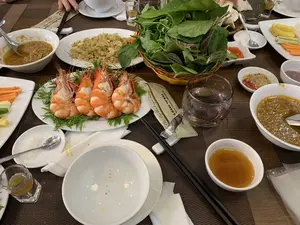 Thông tin văn bản có thể đọc được trên bao bì khăn giấy ướt và khăn giấy: "NHÀ HÀNG VŨ BẢO", "ĐẶC SẢN NGA SƠN", "Chúc Quý khách ngon miệng。", "Hotline: 0886 809 666".。Bối cảnh là một bữa ăn tại một nhà hàng. Mọi người đang cùng nhau thưởng thức các món ăn. Trên bàn có rất nhiều món ăn Việt Nam, nổi bật là một đĩa tôm luộc/hấp lớn được bày biện đẹp mắt, một đĩa chả cá rang sả, một rổ rau sống gồm nhiều loại lá, và một đĩa rau củ quả thái lát (cà rốt, dưa chuột, dứa) để ăn kèm. Ngoài ra còn có nhiều bát nước chấm khác nhau, bao gồm một bát mắm đặc, bát nước mắm ớt, và muối tiêu. Bàn ăn được bày biện với bát, đĩa, đũa, thìa, ly uống nước và ly nhỏ.。（日本語訳）。ウェットティッシュとティッシュのパッケージに記載されている読み取れる文字情報：「VU BAOレストラン」、「NGA SON特産」、「お客様、どうぞ美味しくお召し上がりください。」、「ホットライン：0886 809 666」。背景はレストランでの食事です。人々が一緒に食事を楽しんでいます。テーブルの上にはたくさんのベトナム料理があり、中でも目立つのは、美しく盛り付けられた茹で/蒸しエビの大皿、レモングラスと炒めた魚のすり身揚げの皿、数種類の葉物が入った生野菜のかご、そして付け合わせ用のスライスされた野菜と果物（人参、きゅうり、パイナップル）の皿です。その他にも、様々な種類のつけダレの小鉢があり、濃厚なマム（発酵魚醤）の小鉢、唐辛子入りヌクマム（魚醤）の小鉢、そして塩胡椒が含まれています。食卓には、お椀、皿、箸、スプーン、水用のグラスと小さなグラスが並べられています。