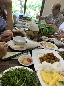 Bức ảnh ghi lại cảnh một bữa ăn đông người tại một nhà hàng, có thể là một buổi sum họp gia đình hoặc bạn bè. Mọi người đang quây quần quanh một bàn ăn thịnh soạn và cùng nhau thưởng thức các món ăn. Hoạt động chính là ăn uống, cụ thể là cuốn các nguyên liệu vào bánh tráng, một phong cách ăn uống đặc trưng của Việt Nam. Trên bàn bày biện rất nhiều món ăn, nổi bật là một đĩa lớn gồm chả cá rán, bún tươi, dứa thái lát, dưa chuột và cà rốt. Xung quanh là các rổ rau thơm, bánh tráng, bánh đa, và nhiều loại nước chấm khác nhau được đặt trong các bát nhỏ, có cả ớt tươi. Các vật dụng trên bàn bao gồm bát sứ trắng, đũa, cốc thủy tinh, và một lon nước ngọt Pepsi ở phía sau. Có thể đọc được dòng chữ trên vỏ bọc đũa: 'Chúc Quý khách ngon miệng' và tên nhà hàng 'NHÀ HÀNG VŨ BẢO' cùng số 'Hotline'. Hộp đựng giấy ăn cũng có logo của nhà hàng.。（日本語訳）。写真は、レストランでの大人数の食事風景を記録したもので、家族や友人の集まりのようです。皆が豪華な食卓を囲み、一緒に料理を楽しんでいます。主な活動は食事で、具体的にはベトナム特有の食事スタイルである、ライスペーパーに具材を巻くことです。テーブルにはたくさんの料理が並べられており、ひときわ目立つのは、揚げさつま揚げ、生のブン（米麺）、スライスしたパイナップル、きゅうり、にんじんが乗った大皿です。周りには、香草の入ったかご、ライスペーパー、バインダー（ライスペーパーを焼いたもの）、そして新鮮な唐辛子も入った様々な種類のつけダレが小鉢に置かれています。テーブルの上にある物には、白い陶器の碗、箸、ガラスのコップ、そして後ろにはペプシの炭酸飲料の缶があります。箸袋には「お客様、どうぞ美味しく召し上がってください」という文字と、「NHÀ HÀNG VŨ BẢO」というレストラン名、そして「ホットライン」の番号が読み取れます。ティッシュボックスにもレストランのロゴが入っています。