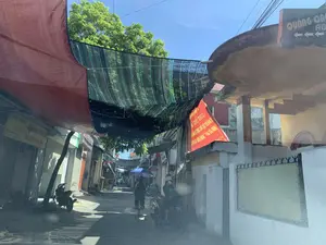 背景は、晴れた日の小さく狭い路地です。路地の上には日よけのために青い防水シートとネットが張られています。両側には家や店がびっしりと並んでいます。**文字：**。1. 右上の高いところにある黒い看板：「QUẢNG CÁO AB...」と書かれ、左を指す3つの矢印が添えられています。2. 左側にある店の黄色い看板：「GIA DỤNG LINH BAN. 0986 580 747. - ĐỒ DÙNG, TIỆN ÍCH. - ĐỒ CHƠI TRẺ EM.」。3. 右側の建物に掲げられた大きな赤い横断幕、読み取れる内容の一部は：「...A KIẾN THỨC」、「...SÁCH VÀ VĂN HÓA ĐỌC...」、「...TẠI XÃ NGƯ LỘC...」です。**背景と活動：**。- **場所の種類：**住宅街または商店のある地区の小さな路地。- **具体的な活動：**日常生活の光景。一人の人が路地で自転車に乗り、日よけに傘をさしています。別の人が右側でバイクに座っています。左側の路肩にはもう一台のバイクが停まっています。- **写真の中の物体：**家々、シャッター付きの店、バイク、自転車、傘、看板、横断幕、日よけの防水シートとネット、緑の植物、そして電線