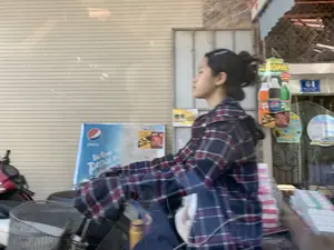 Bức ảnh chụp một cảnh đường phố, có vẻ như được chụp từ một phương tiện đang di chuyển, gây ra hiệu ứng mờ nhòe. Nhân vật chính là một cô gái trẻ đang ngồi trên xe máy, mặc áo sơ mi kẻ sọc màu xanh và đỏ. Tóc cô được búi cao. Phía sau cô là một cửa hàng hoặc nhà dân với cửa cuốn màu be đang đóng. Có một số biển quảng cáo và biển hiệu có thể nhận dạng được: một biển số nhà màu xanh dương ghi số "64", một tấm biển quảng cáo của Pepsi bị mờ, và một biển quảng cáo khác cho các loại nước giải khát (Pepsi, Mirinda, 7 Up) với dòng chữ "TẶNG 250ML". Ngoài ra, có thể thấy một phần của một chiếc xe máy khác màu đỏ và một chiếc giỏ xe ở phía trước.。（日本語訳）。写真は街の風景を撮影したもので、移動中の乗り物から撮影されたようで、ブレ効果が生じています。主な被写体はバイクに座っている若い女性で、青と赤のストライプ柄のシャツを着ています。髪は高く結い上げられています。彼女の後ろには、閉まっているベージュ色のシャッターがある店または民家があります。いくつかの認識可能な広告看板や標識があります：青い住居表示板に「64」という数字、ぼやけたペプシの広告看板、そして「TẶNG 250ML」という文字が書かれた各種清涼飲料水（ペプシ、ミリンダ、セブンアップ）の別の広告看板です。さらに、別の赤いバイクの一部と前方のバイクのかごが見えます。