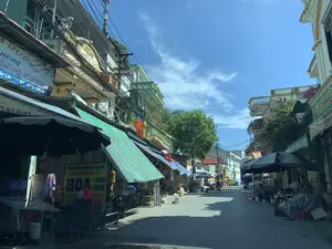 背景は、ある住宅街の小さな通りで、おそらくグーロック（Ngư Lộc）の地元の市場です。空は青く晴れていますが、通りは高層ビルに遮られているため、大部分が日陰になっています。通りの両側には、密集した多層階のチューブハウス（縦長の家）が立ち並び、1階部分には店舗が入っています。店舗は、商品や歩道を日差しから守るために、可動式の軒先テント（緑と白のストライプ、青と白のストライプ）や大きな黒い傘を使用しています。いくつかのベトナム国旗が建物に掲げられています。電線や通信ケーブルが、電柱と家々の間に複雑に張り巡らされています。通りはかなり閑散としており、生活している住民が数人、遠くには自転車に乗る人、そして軒先テントの下に座っている人が数人います。看板で認識できる文字は以下の通りです：。- 左側の青と白の看板：「...H DƯỠNG」、「...ương」、「...IỀM VUI CỦA MẸ」（お母さんの喜び）、「...âu Lộc *** ĐT: 0978.189.283」。これはベビー用品・おむつ店の可能性があります。- 左側の黄色の看板：「Cửa hàng: Đường Nguyệt」（店舗：ドゥオン・グエット）、「CHUYÊN BÁN SỈ - LẺ」（卸売・小売専門）、「HOA - TƯƠI - KHÔ」（花 - 生花 - ドライフラワー）、「NHẬN KẾT HOA... CƯỚI HỎI」（花束の注文受付...結婚式・婚約式）。これは花屋です。- ある建物のファサードには「2010」という数字が刻まれています。- 右手奥の緑色の看板は部分的に読み取ることができ、薬局のようです