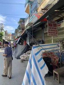 写真はベトナムのある住宅街の通りを撮影したもので、地元の市場のようです。道の両脇には、多層階建てのチューブハウスが密集しています。歩道は仮設の屋台を設置するために使われており、防水シートで覆われています。前景には青と白の縞模様の防水シートが目立ちます。いくつかの店はシャッターを閉めています。帽子をかぶり、縞模様のシャツを着た年配の男性が道端に立っています。奥の方には、数人の人々と一台のバイクが見え、日常の生活風景を作り出しています。バルコニーにはベトナムの金星紅旗が掲げられています。多くの看板が見られ、その中には以下のような内容が記録されています。- ある花屋の赤い広告看板：「生花、各種イベント、フルーツバスケット、生花・ドライフラワーの花輪を専門に取り扱っています。電話：0398.226.591」。- 上部の看板には「NHẤT TÂN」、「TẠO」の文字と2つの電話番号「948352289」、「915594978」があります。- シャッターに貼られた一枚の通知には、「タインホア省人民委員会、保健局、番号：010930.TH.CCHN」という内容が書かれています
