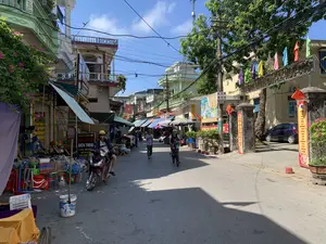 背景は、晴れた日のグーロク村の道です。この道は、両側に店や民家が立ち並ぶ地元の市場です。通りは、バイクや自転車で行き来する人々でかなり賑わっています。左側には、バケツ、たらい、かご、モップなどのプラスチック製の家庭用品を多く販売している店があります。その近くには、「SƠN HIỀN MOBILE 2 携帯電話分割払い販売、ガラス圧着 - 画面交換、バッテリー即時交換」という看板を掲げた携帯電話店と、「VPBank 現金ローン支援」という広告看板があります。右側には、何かのイベントを記念して装飾された公共の建物への入り口があり、おそらく人民委員会の庁舎でしょう。門の上のメインの看板には「...グーロク村人民委員会」と書かれています。門の両側には赤い横断幕が掲げられており、片方には「グーロク村党委員会設立70周年記念を祝う」、もう片方には「デジタル経済、デジタル社会、デジタル市民を目指すことは、4.0時代における必然的なトレンド」と書かれています。門の上には色とりどりの旗がたくさん掲げられています