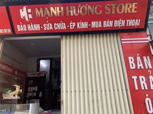 これは「Mạnh Hương Store」という名前の携帯電話店の正面の写真です。店にはクリーム色の金属製シャッターがあり、半分開いています。店のメインの看板は背景が赤色で、「MẠNH HƯƠNG STORE」の文字はブロンズゴールド色、「MH」のロゴがあります。その下には提供されているサービスが白い文字で書かれています：「保証 - 修理 - ガラス圧着 - 携帯電話売買」。電話番号「0923 883 559」も看板に書かれています。右側には「分割払い販売」と書かれた赤い縦長の看板があり、一部が隠れています。店の中を見ると、ガラスのショーケースがあり、その上には装飾として黄色いヤギの像が置かれています。後ろには箱に入った多くの携帯電話アクセサリーを収めた商品棚があり、「SAMSUNG」、「iphone」、「HOCO」などのブランドが見えます