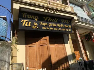 「Trang Nail-Mi」という名前の美容院の正面を撮影した写真です。黄色い立体文字が書かれた大きな黒い看板には、「Trang Nail-Mi」、「Tít」、「シャンプー・ヘアスチーム・修復専門」、「生徒育成 - 電話番号: 0971 612 423」という情報が記載されています。これは、この店がネイルサービス、ヘアケアを専門とし、職業訓練も行っていることを示しています。建物は黄色に塗られており、茶色い木製の正面ドアがあります。左側には古い壁があり、金網のフェンスで保護された青い水タンクがあります。前面には低い白い杭の柵があります。背景は晴天で、空は澄んだ青色です