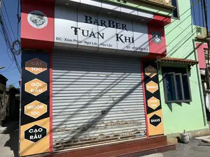 これは「BARBER TUẤN KHỈ」という名前の男性用理髪店の正面の写真です。店は閉まっており、灰色の金属製シャッターが完全に下ろされ、その上にはいくつかの錆の跡があります。ドアの上にあるメインの看板には、店名「BARBER TUẤN KHỈ」、住所「ĐC: Xóm Phúc - Ngư Lộc - Hậu Lộc」、電話番号「ĐT: 0762 800 874」という情報が明記されています。ドアの両側には、ベトナム語と英語の両方でサービスをリストアップした縦長の看板があります。左側の看板には「CẮT TÓC」、「GỘI ĐẦU」、「NHUỘM TÓC」、「CẠO RÂU」と書かれています。右側の看板には「HAIR CUT」、「SHAMPOO」、「HAIR DYE」、「SHAVE」と書かれています。さらに、正面には2つの円形のロゴがあります。一方のロゴには「BARBERSHOP」、「THE BEST」、「SHAVE & HAIRCUT」、「PARLOR」と書かれており、もう一方のロゴには店名「TUẤN KHỈ」、「BARBER SHOP」と設立年「EST 2019」が書かれています。店の建物は赤と白の正面をしており、緑色に塗られた家の隣にあります。背景は晴れた日で、空は青く澄んでいます