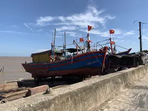 Bức ảnh chụp một chiếc tàu đánh cá bằng gỗ đang được kéo lên bờ tại một khu vực ven biển, có thể là một cảng cá hoặc bãi sửa chữa tàu thuyền. Thân tàu có ghi dòng chữ 'TH 90541 TS'. Chiếc tàu có kích thước lớn, vỏ gỗ được sơn màu xanh và đỏ, cabin màu vàng, trông đã khá cũ. Tàu đang nằm trên một hệ thống triền đà (đường ray để kéo tàu lên bờ) bên cạnh một bờ kè bê tông. Trên các cột buồm của tàu treo nhiều lá cờ Tổ quốc Việt Nam (cờ đỏ sao vàng). Khung cảnh cho thấy con tàu đang trong thời gian neo đậu để sửa chữa hoặc bảo dưỡng. Phía sau là vùng nước rộng lớn của biển hoặc cửa sông, nước có màu nâu phù sa. Xa xa trên mặt nước có thể nhìn thấy nhiều chòi canh, đây là hình ảnh quen thuộc của các khu vực nuôi trồng thủy sản (như nuôi ngao). Bầu trời trong xanh, có nắng và mây trắng.。（日本語訳）。写真は、木造の漁船が沿岸地域、おそらく漁港か船舶修理場で陸に引き上げられているところを写しています。船体には「TH 90541 TS」という文字が書かれています。船は大型で、木製の船体は青と赤に塗られ、船室は黄色で、かなり古びて見えます。船は、コンクリートの護岸の隣にある船台（船を陸に引き上げるためのレール）の上にあります。船のマストには、多くのベトナム国旗（金星紅旗）が掲げられています。この光景は、船が修理またはメンテナンスのために停泊中であることを示しています。後方には海または河口の広大な水域が広がり、水は沖積土で茶色く濁っています。遠くの水面には多くの見張り小屋が見え、これは（アサリの養殖などの）水産養殖地域でよく見られる光景です。空は青く晴れ渡り、日差しがあり、白い雲が浮かんでいます。