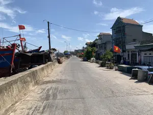 Bối cảnh là một con đường ven biển tại một làng chài vào một ngày nắng đẹp, trời trong xanh. Bên trái là một bờ kè bê tông, nơi các tàu đánh cá bằng gỗ lớn, sơn màu xanh và đỏ, đang neo đậu. Trên cột buồm của tàu có cắm cờ Việt Nam. Có thể nhìn thấy một phần ký tự '...HTS' trên thân một chiếc tàu, có thể là số hiệu tàu. Dọc theo bờ kè còn có một số lán trại tạm. Ở giữa là con đường nhựa khá vắng vẻ. Bên phải là dãy nhà dân nhiều tầng, một số nhà có treo cờ Việt Nam ở phía trước. Có một vài người dân đang ngồi nghỉ và một vài chiếc xe máy đậu ven đường. Khung cảnh chung cho thấy cuộc sống sinh hoạt gắn liền với nghề đi biển.。（日本語訳）。背景は、晴れた美しい日、空が青く澄んだ漁村の海岸沿いの道です。左側はコンクリートの護岸で、そこには青と赤に塗られた大きな木造の漁船が停泊しています。船のマストにはベトナムの国旗が掲げられています。一隻の船の船体には「...HTS」という文字の一部が見え、それは船の登録番号かもしれません。護岸に沿って、いくつかの仮設の小屋もあります。中央には、かなり閑散としたアスファルトの道があります。右側は多層階の民家が並んでおり、いくつかの家は前面にベトナムの国旗を掲げています。何人かの住民が座って休んでおり、何台かのバイクが道端に停まっています。全体的な光景は、漁業と密接に結びついた日常生活を示しています。