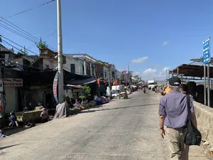 Bản ghi các thông tin bằng chữ:。- Trên biển hiệu của một cửa hàng bên trái có ghi: 'Hùng Huyền BÁN LẺ HẢI SẢN TƯƠI SỐNG ĐT: 0972 248 404 - 0983 832 067'.。- Trên biển báo màu xanh dựng bên phải đường có ghi: 'XÃ NGƯ LỘC ĐOẠN ĐƯỜNG ĐÊ TỰ QUẢN VỀ VỆ SINH MÔI TRƯỜNG'.。Mô tả cảnh quan và hoạt động:。Hình ảnh ghi lại khung cảnh sinh hoạt trên một con đường bê tông tại một làng chài ven biển, được xác định là xã Ngư Lộc qua thông tin trên biển báo. Con đường này là một đoạn đường đê, một bên là dãy nhà ở kết hợp kinh doanh và bên còn lại có thể là khu vực bến cảng.。Dọc theo con đường là các hoạt động buôn bán tấp nập, đặc biệt là hải sản tươi sống như trên biển hiệu. Người dân đang di chuyển bằng nhiều phương tiện: một người đàn ông lớn tuổi đang đi bộ, một người phụ nữ đẩy xe kéo ba bánh, và một người khác đi xe máy. Một số người đội nón lá đang làm việc hoặc mua bán trước các cửa hàng. Các vật thể trong ảnh bao gồm nhà cửa, cột điện với dây điện chằng chịt, xe tải, cờ Việt Nam treo trước một số nhà, và các hàng hóa được che đậy bằng bạt.。（日本語訳）。文字情報の記録：。- 左側の店の看板には「フン・フエン 生鮮海産物小売 電話：0972 248 404 - 0983 832 067」と書かれている。- 道路の右側に立てられた青い看板には「グーロック社 環境衛生自主管理堤防道路区間」と書かれている。景観と活動の描写：。写真は、看板の情報からグーロック社と特定される、沿岸の漁村にあるコンクリート道路での生活風景を記録している。この道は堤防道路の一部で、片側には住居兼店舗が並び、もう片側は港湾地区である可能性がある。道沿いでは、看板にあるような生鮮海産物を中心に、活発な商売活動が行われている。人々は様々な交通手段で移動している：年配の男性が歩いており、女性が三輪の荷車を押し、別の人がバイクに乗っている。何人かの人々がノンラー（菅笠）をかぶり、店の前で働いたり、売買をしたりしている。写真の中の物体には、家々、絡み合った電線のある電柱、トラック、いくつかの家の前に掲げられたベトナム国旗、そして防水シートで覆われた商品などが含まれる。