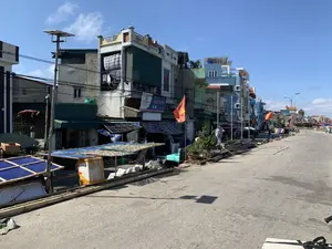 Bối cảnh là một con đường bê tông rộng rãi ven biển tại một làng chài vào một ngày nắng, trời trong xanh. Dọc theo con đường là một dãy nhà ống nhiều tầng, được xây san sát nhau với nhiều màu sắc và kiến trúc đa dạng. Hoạt động chính diễn ra là phơi hải sản; cá được trải trên các tấm liếp lớn đặt trên những giàn phơi tạm bợ làm bằng tre ngay bên lề đường. Nhiều lá cờ đỏ sao vàng của Việt Nam được treo trước các ngôi nhà. Có một vài người dân đang sinh hoạt và làm việc bên đường. Các vật dụng khác có thể thấy bao gồm các cây tre, luồng dài được xếp chồng lên nhau, một chiếc xe máy đang đỗ, và các cột đèn, trong đó có một cột đèn sử dụng tấm pin năng lượng mặt trời. Một tấm biển hiệu có thể đọc được ghi chữ 'HẢI THỦY' và logo của 'SIAM Brothers'.。（日本語訳）。背景は、ある晴れた日の漁村にある、広々とした海沿いのコンクリート道路で、空は青く澄んでいる。道に沿って、多種多様な色と建築様式の多層階建てのチューブハウスが密集して建ち並んでいる。主な活動は海産物を干すことで、魚が道端のすぐそばにある竹製の簡易的な干し台の上に置かれた大きなすのこの上に広げられている。何人かの住民が道端で生活したり仕事をしたりしている。他に見られる物には、積み重ねられた長い竹やルオン、駐車している一台のバイク、そして街灯が含まれ、その中にはソーラーパネルを使用している街灯が一本ある。「HẢI THỦY」という文字と「SIAM Brothers」のロゴが書かれた、読み取れる看板が一つある。