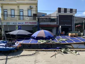 Bức ảnh chụp một khung cảnh đường phố với các hoạt động buôn bán. Ở hậu cảnh là những ngôi nhà phố nhiều tầng. Một trong số đó có biển hiệu lớn ghi 'ĐẠI LÝ SỸ THÔNG', với thông tin 'ĐỊA CHỈ: NGƯ LỘC - HẬU LỘC - THANH HÓA' và 'ĐT: 0974 487 748'. Các biển hiệu khác như 'RYX LUBRICANTS' và 'PINACO' cho thấy đây là một đại lý kinh doanh dầu nhớt và ắc quy. Phía trước, trên mặt đường bê tông, có các sạp hàng tạm được dựng bằng khung tre và lợp bạt nhựa màu xanh. Hai chiếc ô lớn được mở để che nắng cho khu vực bán hàng. Có một vài người đang ngồi dưới các sạp hàng, trong đó có một người đội nón lá. Xung quanh có các vật dụng như rổ nhựa, thùng carton và các tấm bạt. Cảnh tượng cho thấy một khu chợ tạm hoặc các quầy hàng ven đường vào một ngày nắng.。（日本語訳）。写真は、商売が行われている街頭の風景を写しています。背景には、複数階建てのタウンハウスがあります。そのうちの一つには、「ĐẠI LÝ SỸ THÔNG」と書かれた大きな看板があり、「ĐỊA CHỈ: NGƯ LỘC - HẬU LỘC - THANH HÓA」（住所：グーロック - ハウロック - タインホア）と「ĐT: 0974 487 748」（電話：0974 487 748）という情報が記載されています。「RYX LUBRICANTS」や「PINACO」といった他の看板は、ここが潤滑油とバッテリーを扱う代理店であることを示しています。手前のコンクリートの路上には、竹の骨組みで建てられ、青いビニールシートで覆われた仮設の屋台があります。売り場の日よけのために、2つの大きな傘が開かれています。屋台の下には数人が座っており、その中にはノンラーをかぶった人が一人います。周りには、プラスチック製のカゴ、段ボール箱、シートなどの物があります。この光景は、晴れた日の仮設市場または道端の露店を示しています。