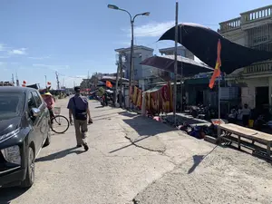 Bối cảnh là một con đường bê tông vào một ngày nắng ở một làng chài ven biển, có thể là khu vực cảng cá. Một người đàn ông đang đi bộ trên đường, tay cầm máy ảnh. Gần đó, một người phụ nữ đội nón lá đang đứng cạnh chiếc xe đạp của mình, bên cạnh một chiếc ô tô màu đen đang đỗ. Dọc theo con đường là những ngôi nhà cao tầng và các hoạt động sinh hoạt của người dân. Có những người đang ngồi bên lề đường, có thể đang bán hàng hoặc làm việc, bên cạnh một chiếc bàn gỗ. Nhiều xe máy đang được đỗ. Nhiều lá cờ Tổ quốc Việt Nam và cờ Đảng được treo dọc đường và trên các tàu thuyền đánh cá ở phía xa. Chăn mền được phơi trên dây. Có các tấm bạt lớn được căng ra để che nắng. Một tấm biển hiệu có ghi chữ 'TÂM ANH' nhưng các chi tiết khác không thể đọc được.。（日本語訳）。背景は、沿岸の漁村にある晴れた日のコンクリートの道で、おそらく漁港のエリアです。一人の男性が道を歩いており、手にはカメラを持っています。近くでは、ノンラーをかぶった一人の女性が自分の自転車のそばに立っており、その隣には黒い車が駐車しています。道沿いには高層の家々と人々の生活の営みがあります。道端に座っている人々がおり、おそらく商品を売っているか仕事をしており、そのそばには木製のテーブルがあります。多くのバイクが駐車されています。多くのベトナム国旗と党旗が道沿いや遠くの漁船に掲げられています。毛布がロープに干されています。日差しを遮るために大きな防水シートが張られています。「TÂM ANH」という文字が書かれた看板がありますが、他の詳細は読み取れません。