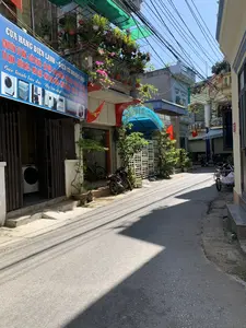 Bối cảnh là một con hẻm nhỏ, có nhiều nhà dân san sát nhau vào một ngày nắng. Bên trái là một cửa hàng sửa chữa và kinh doanh đồ điện lạnh, điện tử có tên 'HOÀNG ĐẠI'. Tấm biển hiệu lớn màu xanh của cửa hàng ghi rõ các dịch vụ và thông tin liên lạc: 'CỬA HÀNG ĐIỆN LẠNH - ĐIỆN TỬ HOÀNG ĐẠI', 'CHUYÊN TƯ VẤN - SỬA CHỮA - LẮP ĐẶT - BẢO DƯỠNG - MUA BÁN - TRAO ĐỔI CŨ - MỚI', 'TỦ LẠNH - ĐIỀU HÒA - MÁY GIẶT - MÁY LỌC NƯỚC - MÁY RỬA BÁT - BÌNH NÓNG LẠNH - BẾP TỪ', và 'Bảo hành lâu dài - Uy tín chất lượng ĐT: 0972.956.526'. Cửa xếp của cửa hàng đang hé mở, có thể thấy một chiếc máy giặt bên trong. Phía trước một số ngôi nhà có treo cờ Tổ quốc Việt Nam (cờ đỏ sao vàng). Ban công của ngôi nhà có cửa hàng được trồng rất nhiều chậu cây cảnh và hoa giấy. Một chiếc xe máy đang đỗ trên vỉa hè gần cửa hàng. Nhìn sâu vào trong hẻm, có thể thấy thêm vài chiếc xe máy và một chiếc xe ba gác. Phía trên là hệ thống dây điện chằng chịt, một hình ảnh quen thuộc tại các khu dân cư ở Việt Nam.。（日本語訳）。舞台は晴れた日の小さな路地で、多くの民家が密集している。左側には「HOÀNG ĐẠI」という名前の、冷凍・電子機器の修理・販売店がある。店の大きな青い看板には、サービス内容と連絡先が明記されている。「冷凍・電子機器店 HOÀNG ĐẠI」、「専門：コンサルティング - 修理 - 設置 - メンテナンス - 売買 - 中古・新品交換」、「冷蔵庫 - エアコン - 洗濯機 - 浄水器 - 食器洗い機 - 給湯器 - IHコンロ」、そして「長期保証 - 信頼と品質 電話：0972.956.526」。店の近くの歩道にバイクが1台停まっている。上には複雑に絡み合った電線があり、ベトナムの住宅地ではおなじみの光景である。