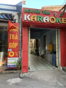 Hình ảnh chụp mặt tiền của một quán karaoke có tên 'NGUYÊN TÂM KARAOKE'. Biển hiệu chính của quán có nền đỏ, chữ 'NGUYÊN TÂM' màu xanh lá cây và chữ 'KARAOKE' nhiều màu sắc (trắng, vàng, xanh lá), các chữ cái được gắn đèn LED. Lối vào quán mở ra một con hẻm nhỏ, hẹp, nền được lát gạch hoa. Bên trong con hẻm có thể thấy một chiếc ghế nhựa màu xanh, thùng xốp, thùng phuy nhựa màu xanh và các vật dụng khác. Bên trái là một cửa hàng khác có treo lá cờ Việt Nam và một biển quảng cáo dọc màu đỏ với chữ vàng ghi 'BÁN TRẢ GÓP 0%'. Phía dưới biển quảng cáo này có thông tin địa chỉ và số điện thoại: 'ĐC: THẮNG LỘC - NG...', 'ĐT: 0965348...'. Khung cảnh là một con phố với các cửa hàng, không có hoạt động nào của con người được ghi lại.。（日本語訳）。「NGUYÊN TÂM KARAOKE」という名前のカラオケ店の正面を撮影した写真。店のメイン看板は赤地で、「NGUYÊN TÂM」の文字は緑色、「KARAOKE」の文字は多色（白、黄、緑）で、文字にはLEDライトが取り付けられている。店の入り口は狭い小さな路地に通じており、床には模様付きのタイルが敷かれている。この広告看板の下には、住所と電話番号の情報がある：「ĐC: THẮNG LỘC - NG...」、「ĐT: 0965348...」。風景は店舗が並ぶ通りで、人の活動は記録されていない。