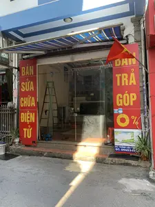 Hình ảnh chụp mặt tiền của một cửa hàng ven đường, có vẻ là một cửa hàng sửa chữa và bán đồ điện tử. Cửa hàng có cửa trượt bằng kính và mái hiên sọc xanh trắng. Ở hai bên lối vào là các biển hiệu dọc màu đỏ với chữ vàng. Biển hiệu bên trái ghi 'BÁN SỬA CHỮA ĐIỆN TỬ'. Biển hiệu bên phải ghi 'BÁN TRẢ GÓP 0%'. Một lá cờ Việt Nam (cờ đỏ sao vàng) được cắm ở phía trước. Dưới biển hiệu bên phải có một tấm áp phích quảng cáo TV với các thông tin: 'TIVI 43 INCH SMART', 'ĐC: THẮNG LỘC - NGƯ', và 'ĐT: 0965348...'. Bên trong cửa hàng, có thể nhìn thấy một chiếc thang chữ A. Khung cảnh tĩnh, không có người và cửa hàng dường như đang đóng cửa. Xung quanh có một vài chậu cây cảnh, một chiếc ghế nhựa màu đỏ và đường phố phía trước có ánh nắng chiếu rọi.。（日本語訳）。道端にある店の正面を撮影した写真で、電子機器の修理・販売店のようです。店にはガラスの引き戸と、青と白の縞模様の庇があります。入り口の両側には、黄色い文字の入った赤い縦長の看板があります。左側の看板には「BÁN SỬA CHỮA ĐIỆN TỬ」と書かれています。右側の看板には「BÁN TRẢ GÓP 0%」と書かれています。店の前にはベトナム国旗（金星紅旗）が掲げられています。右側の看板の下にはテレビの広告ポスターがあり、「TIVI 43 INCH SMART」、「ĐC: THẮNG LỘC - NGƯ」、「ĐT: 0965348...」という情報が記載されています。店内には、A字型のはしごが見えます。静かな光景で、人はおらず、店は閉まっているようです。周りにはいくつかの観葉植物の鉢、赤いプラスチックの椅子があり、前の通りには日差しが差し込んでいます。