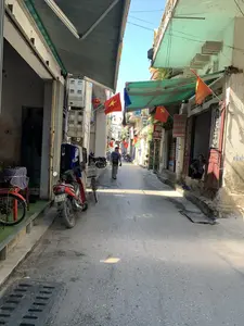 Bối cảnh là một con ngõ nhỏ và hẹp tại một khu dân cư ở Việt Nam vào một ngày nắng. Hai bên ngõ là những ngôi nhà san sát nhau, một số nhà được sử dụng làm cửa hàng kinh doanh. Dọc theo con ngõ, nhiều lá cờ Tổ quốc (cờ đỏ sao vàng) được treo lên, cho thấy không khí của một ngày lễ hoặc sự kiện quốc gia. Một người đàn ông đang đi bộ sâu vào trong ngõ. Ở cửa một ngôi nhà bên phải, một người phụ nữ lớn tuổi đang ngồi và nhìn ra ngoài. Nhiều xe máy và xe đạp được đỗ dọc hai bên lối đi. Các cửa hàng có mái hiên di động để che nắng. Một cửa hàng bên trái treo quần áo để bán. Có thể nhìn thấy các biển hiệu với nội dung: 'TẠI ĐÂY Chuyên SỬA CHỮA QUẦN ÁO QUẦN BÒ 0966419784' và một biển hiệu khác có chữ 'SỬA ĐÔNG H...'. Trên tường bên phải có dòng chữ viết bằng sơn đã mờ: 'KNCB 0... 267...'.。（日本語訳）。背景は、ある晴れた日のベトナムの住宅街にある、小さく狭い路地です。路地の両側には家々が密集しており、そのうちのいくつかは商店として使われています。路地に沿って、多くの国旗（金星紅旗）が掲げられており、祝日か国家的なイベントの雰囲気を示しています。一人の男性が路地の奥へと歩いています。右側にある家の戸口では、年配の女性が座って外を眺めています。通路の両脇に沿って、多くのバイクや自転車が停められています。商店には日差しを遮るための可動式の日よけがあります。左側の店は販売用の服を吊るしています。「TẠI ĐÂY Chuyên SỬA CHỮA QUẦN ÁO QUẦN BÒ 0966419784」という内容の看板と、「SỬA ĐÔNG H...」という文字がある別の看板が見えます。右側の壁には、「KNCB 0... 267...」という、色褪せたペンキで書かれた文字があります。