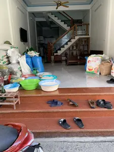 Bối cảnh là không gian mở phía trước của một ngôi nhà, đồng thời cũng là một cửa hàng kinh doanh. Nhiều bao tải lớn được xếp chồng lên nhau, trong đó có các bao gạo và có thể là thức ăn chăn nuôi. Một bao gạo trắng có dòng chữ 'ĐẶC SẢN Đ... HẠT NGỌC VÙNG CAO NGUYÊN ST 25' và '50kg'. Một bao khác có thương hiệu 'Cargill'. Trên bậc thềm dẫn vào nhà, nhiều chậu nhựa màu sắc khác nhau (xanh lá, xanh dương) của thương hiệu 'VIETNHAT' được dùng để đựng gạo bán lẻ, bên cạnh có một chiếc cân đồng hồ. Bên trong nhà có thể thấy nội thất gia đình như bộ bàn ghế gỗ, cầu thang lên lầu với lan can gỗ và kính, và một chiếc TV treo tường. Phía trước có một chiếc xe máy màu đỏ đang đậu. Nhiều đôi dép được để trên bậc thềm. Khung cảnh cho thấy đây là một cửa hàng bán gạo và nông sản tại gia.。（日本語訳）。背景は、ある家の前のオープンスペースであり、同時に商店でもある。大きな袋がいくつも積み重ねられており、その中には米袋や、おそらくは飼料も含まれている。ある白い米袋には「ĐẶC SẢN Đ... HẠT NGỌC VÙNG CAO NGUYÊN ST 25」と「50kg」という文字がある。別の袋には「Cargill」というブランド名がある。家に通じる上がり口には、「VIETNHAT」ブランドの様々な色（緑、青）のプラスチック製の鉢がいくつも置かれ、米を小分けにして販売するために使われており、その隣にはアナログの秤がある。前方には赤いバイクが1台停まっている。この光景は、ここが自宅で米や農産物を販売する店であることを示している。