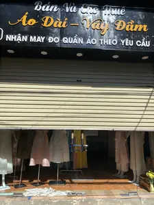 Bức ảnh chụp mặt tiền của một cửa hàng thời trang. Biển hiệu màu đen ở phía trên có ghi "Bán Và Cho Thuê Áo Dài - Váy Đầm" và "NHẬN MAY ĐO QUẦN ÁO THEO YÊU CẦU". Cửa hàng có cửa cuốn bằng kim loại màu be, được kéo lên một phần, để lộ không gian trưng bày bên trong qua lớp cửa kính. Bên trong, có nhiều ma-nơ-canh đang mặc các mẫu áo dài và váy với màu sắc khác nhau. Sàn nhà được lát gạch men hoa văn. Cửa hàng có vẻ đang đóng cửa hoặc chưa mở cửa hoàn toàn.。（日本語訳）。ファッション店の正面を撮影した写真です。上部にある黒い看板には「アオザイ・ドレスの販売とレンタル」そして「オーダーメイド服の受注」と書かれています。店にはベージュ色の金属製シャッターがあり、一部が引き上げられており、ガラス戸越しに中の陳列スペースが見えています。中には、様々な色のアオザイやドレスを着た多くのマネキンがあります。床は模様入りのセラミックタイルで敷き詰められています。店は閉まっているか、まだ完全には開店していないようです。