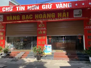 Bức ảnh chụp mặt tiền của một tiệm vàng bạc tên là "VÀNG BẠC HOÀNG HẢI". Cửa hàng có mặt tiền màu đỏ nổi bật, được chụp vào một ngày nắng đẹp.。**Chữ viết được ghi lại:**。*   Biển hiệu trên cùng, chữ trắng trên nền đỏ: "CHỮ TÍN HƠN GIỮ VÀNG".。*   Biển hiệu chính, chữ vàng trên nền đỏ: "VÀNG BẠC HOÀNG HẢI".。*   Thông tin địa chỉ và điện thoại: "ĐC: ĐỐI DIỆN TƯỢNG ĐÀI XÃ NGƯ LỘC", "ĐT: 0986 957 798 - 0343 957 597".。*   Các biển hiệu dọc trên cột: "UY TÍN - CHẤT LƯỢNG", "NHẪN ĐÔI", "NHẪN CƯỚI", "SỬA CHỮA", và các chữ khác như "TRAO ĐỔI", "MUA BÁN".。*   Logo "HH" được lặp lại ở nhiều nơi.。*   Trên cửa kính có dán chỉ dẫn "ĐÂY SANG".。*   Bên trong có treo đèn lồng đỏ với chữ "TIỀN VÀO NHƯ NƯỚC".。**Mô tả cảnh quan và hoạt động:**。*   **Loại địa điểm:** Đây là một cửa hàng kinh doanh vàng bạc, tọa lạc tại xã Ngư Lộc.。*   **Hoạt động:** Cửa hàng có vẻ đang đóng cửa hoặc chỉ mở một phần, vì cửa cuốn màu nâu được kéo xuống gần hết. Không có hoạt động giao dịch nào rõ ràng diễn ra ở bên ngoài. Bên trong qua lớp cửa kính, có thể thấy lờ mờ bóng người và các tủ trưng bày bằng gỗ.。*   **Vật thể:** Nổi bật là các biển hiệu quảng cáo lớn màu đỏ. Phía trước cửa hàng có cửa cuốn, cửa ra vào bằng kính, bậc tam cấp, một vài chậu cây cảnh, một tấm dốc kim loại nhỏ để dắt xe, và một vài đôi dép để trên bậc thềm.。（日本語訳）。「VÀNG BẠC HOÀNG HẢI」という名前の金銀店の正面を写した写真です。店は目立つ赤色のファサードをしており、晴れた日に撮影されました。**記録された文字：**。*   一番上の看板、赤地に白文字：「CHỮ TÍN HƠN GIỮ VÀNG」（信用は金を保つより尊い）。*   メインの看板、赤地に金文字：「VÀNG BẠC HOÀNG HẢI」（ホアン・ハイ金銀店）。*   住所と電話番号の情報：「ĐC: ĐỐI DIỆN TƯỢNG ĐÀI XÃ NGƯ LỘC」（住所：グーロック社記念碑の向かい）、「ĐT: 0986 957 798 - 0343 957 597」（電話：0986 957 798 - 0343 957 597）。*   柱にある縦長の看板：「UY TÍN - CHẤT LƯỢNG」（信頼 - 品質）、「NHẪN ĐÔI」（ペアリング）、「NHẪN CƯỚI」（結婚指輪）、「SỬA CHỮA」（修理）、及び「TRAO ĐỔI」（交換）、「MUA BÁN」（売買）などの他の文字。*   「HH」のロゴが多くの場所で繰り返されています。*   ガラスのドアには「ĐÂY SANG」（こちらへ）という案内が貼られています。*   店内には「TIỀN VÀO NHƯ NƯỚC」（お金が水のように入ってくる）という文字が書かれた赤い提灯が吊るされています。**風景と活動の描写：**。*   **場所の種類：** これはグーロック社に位置する金銀を商う店です。*   **活動：** 店は閉店しているか、一部のみ営業しているように見えます。なぜなら、茶色のシャッターがほとんど下ろされているからです。外では明確な取引活動は行われていません。ガラスのドア越しに店内を見ると、人影や木製の陳列棚がぼんやりと見えます。*   **物体：** 目立つのは大きな赤い広告看板です。店の前にはシャッター、ガラスの出入り口、3段の階段、いくつかの観葉植物の鉢、バイクを押して上がるための小さな金属製のスロープ、そして階段に置かれたいくつかのサンダルがあります。