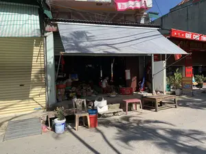 Bối cảnh là một con phố vào một ngày nắng. Ở trung tâm bức ảnh là một cửa hàng tạp hóa đang mở cửa, được che bằng một tấm bạt lớn màu xám để tránh nắng. Bên trong cửa hàng, một người phụ nữ đeo khẩu trang đang ngồi trên một chiếc ghế nhựa đỏ, dường như đang sơ chế một loại củ quả nào đó trong một chiếc chậu kim loại. Cửa hàng bày bán nhiều loại hàng hóa khác nhau, có thể thấy các thùng hàng, chai nước ngọt, một chiếc cân và các vật dụng khác. Phía trước cửa hàng, trên vỉa hè, có đặt một chiếc ghế nhựa, một chiếc bàn gỗ nhỏ, vài cái xô và chậu cây.。Ngôi nhà bên trái có cửa cuốn màu vàng đang đóng, phía trên là mái hiên sọc xanh trắng. Ngôi nhà bên phải có một biển hiệu màu đỏ và một tấm biển quảng cáo dịch vụ tài chính dựng đứng.。Các dòng chữ có thể đọc được trong ảnh bao gồm:。- Trên biển hiệu màu đỏ bên phải: "* * * GIỮ CHỮ TÍN", "...NG BẠC HOÀNG H...", "UY TÍN - CHẤT LƯỢNG".。- Trên biển quảng cáo màu vàng: "NHẬN CHUYỂN TIỀN", "RÚT TIỀN MẶT", "QUẸT THẺ MỌI NGÂN HÀNG", "THU HỘ TRẢ GÓP", "CHI PHÍ THẤP", và số điện thoại "098 695 7798".。- Trên cửa cuốn bên trái: "Navidoor".。- Trên chiếc chăn phơi ở ban công tầng trên: "Vijako".。- Một số điện thoại dán trên tường: "...571 939 282".。（日本語訳）。背景は晴れた日のとある通りです。写真の中央には、日差しを避けるために大きな灰色のシートで覆われた、営業中の雑貨店があります。店の中では、マスクをした女性が赤いプラスチックの椅子に座り、金属製のたらいの中で何らかの根菜か果物を下ごしらえしているようです。店には様々な商品が陳列されており、段ボール箱、ソフトドリンクのボトル、はかり、その他の品々が見えます。店の前、歩道の上には、プラスチックの椅子、小さな木製のテーブル、いくつかのバケツ、植木鉢が置かれています。左側の家には閉まっている黄色のシャッターがあり、その上には青と白の縞模様の庇があります。右側の家には赤い看板と、縦置きの金融サービスの広告看板があります。写真で読み取れる文字は以下の通りです。- 右側の赤い看板：「* * * GIỮ CHỮ TÍN（信用を守る）」、「...NG BẠC HOÀNG H...」、「UY TÍN - CHẤT LƯỢNG（信頼 - 品質）」。- 黄色い広告看板：「NHẬN CHUYỂN TIỀN（送金受付）」、「RÚT TIỀN MẶT（現金引き出し）」、「QUẸT THẺ MỌI NGÂN HÀNG（全銀行カード対応）」、「THU HỘ TRẢ GÓP（分割払い代行回収）」、「CHI PHÍ THẤP（低料金）」、そして電話番号「098 695 7798」。- 左側のシャッター：「Navidoor」。- 上階のバルコニーに干されている毛布：「Vijako」。- 壁に貼られた電話番号：「...571 939 282」。