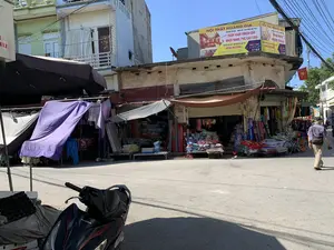 Bức ảnh chụp một con phố buôn bán ở Việt Nam vào một ngày nắng. Trên nóc một tòa nhà có một tấm biển quảng cáo lớn màu vàng với nội dung: "NỘI THẤT HOÀNG GIA. CHUYÊN TƯ VẤN - THIẾT KẾ - THI CÔNG NỘI THẤT. TRẦN VÁCH THẠCH CAO. NHỰA NANO, PVC CÁC LOẠI. CHUYÊN CUNG CẤP, PHÂN PHỐI CÁC LOẠI SƠN JOTUN, MISTASHU,...". Biển hiệu cũng ghi địa chỉ và thông tin liên lạc: "ĐC: Minh Thịnh - Minh Lộc - Hậu Lộc - Thanh Hóa. ĐT: 0978 082 567. FB: Hoang Viet. Zalo: 0978 082 567".。Khung cảnh là một khu phố với các cửa hàng san sát nhau. Các cửa hàng này đang hoạt động, bày bán nhiều loại hàng hóa ra vỉa hè, chủ yếu là đồ gia dụng như đệm, gối, chăn, chiếu cuộn và quần áo. Các gian hàng được che bằng ô và bạt lớn để tránh nắng. Một người đàn ông đang đi bộ trên đường. Một lá cờ Việt Nam được treo trên một tòa nhà. Ở phía trước, một phần của chiếc xe máy hiệu Honda màu đen đang đỗ.。（日本語訳）。写真は、晴れた日のベトナムの商店街を写したものです。ある建物の屋上には、黄色い大きな看板があり、次のような内容が書かれています。「HOANG GIAインテリア。インテリアのコンサルティング・設計・施工専門。石膏ボード天井・壁。各種ナノプラスチック、PVC。JOTUN、MISTASHUなどの各種塗料の供給・販売専門…」。看板には住所と連絡先も記載されています。「住所：タインホア省ハウロック県ミンロック社ミンティン。電話：0978 082 567。FB：Hoang Viet。Zalo：0978 082 567」。景色は、店が隣り合って立ち並ぶ商店街です。これらの店は営業中で、歩道には様々な商品が並べられており、主にマットレス、枕、毛布、巻きござ、衣類などの家庭用品です。各店舗は日差しを避けるために、傘や大きなシートで覆われています。一人の男性が道を歩いています。ある建物にはベトナム国旗が掲げられています。手前には、黒いホンダ製のバイクの一部が停まっています。