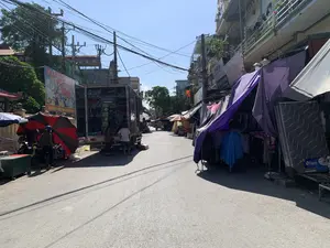 Đây là hình ảnh một con phố chợ ngoài trời ở Việt Nam vào một ngày nắng. Con đường được trải nhựa, hai bên là các tòa nhà san sát và nhiều sạp hàng tạm bợ. Phía trên là hệ thống dây điện chằng chịt. Bên trái đường, một chiếc xe tải lớn đang đỗ và mở thùng sau, bên trong chứa đầy hàng hóa như chăn, đệm được xếp trên kệ. Một vài người đang đứng xem hàng ở đuôi xe tải. Gần đó, có một người đội mũ bảo hiểm đứng cạnh xe máy dưới một chiếc ô lớn màu đỏ và đen. Bên phải đường là một dãy các sạp hàng được che bằng bạt, chủ yếu là bạt màu tím, bày bán quần áo treo trên giá và một chiếc đệm có nhãn hiệu "VIJAKO". Có một tấm áp phích lớn ở bên trái với nội dung "Chúc Mừng QUỐC KHÁNH 2-9" và "TINH THẦN CÁCH MẠNG THÁNG TÁM VÀ QUỐC KHÁNH 2-9 BẤT DIỆT", cùng hình ảnh Chủ tịch Hồ Chí Minh. Trên các tòa nhà bên phải có nhiều biển hiệu cửa hàng nhưng khó đọc rõ, có thể thấy các chữ như "QUẦN ÁO...", "THIÊN LONG", "HUY DINH". Khung cảnh thể hiện hoạt động buôn bán, sinh hoạt thường ngày tại một khu chợ địa phương.。（日本語訳）。これは晴れた日のベトナムの屋外市場の通りの写真です。道路は舗装されており、両側には建物が密集し、多くの仮設の屋台が並んでいます。上には電線が複雑に張り巡らされています。道路の左側には、大きなトラックが停車し、後ろの荷台を開けています。中には棚に並べられた毛布やマットレスなどの商品がいっぱいです。数人がトラックの後ろで商品を眺めています。その近くでは、ヘルメットをかぶった人が赤と黒の大きな傘の下でバイクのそばに立っています。道路の右側には、主に紫色のシートで覆われた屋台が並んでおり、ラックに掛けられた衣類や「VIJAKO」というブランドのマットレスが販売されています。左側には、「9月2日建国記念日おめでとう」と「8月革命と9月2日建国記念日の精神は不滅」という内容の大きなポスターがあり、ホー・チ・ミン主席の写真も描かれています。右側の建物には多くの店の看板がありますが、はっきりと読み取るのは難しく、「QUẦN ÁO...」（衣類...）、「THIÊN LONG」、「HUY DINH」などの文字が見えます。この光景は、地元の市場での日常的な商売や生活の様子を表しています。