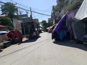 Khung cảnh một con phố nhỏ, đông đúc vào một ngày nắng, có vẻ là một khu chợ đường phố. Hai bên đường là các tòa nhà nhiều tầng san sát, phía trên có hệ thống dây điện chằng chịt. Bên trái, một chiếc xe tải lớn đang đỗ với thùng xe mở, có vài người đang đứng xung quanh, dường như đang bốc dỡ hàng hóa là chiếu và đệm. Gần đó, một quầy hàng nhỏ được che bởi một chiếc ô lớn màu đỏ và đen. Bên phải đường, các quầy hàng tạm được dựng lên, che bằng bạt màu tím và trắng, bày bán các mặt hàng như quần áo và đệm. Một chiếc đệm có nhãn hiệu "VIJAKO" được dựng lên để trưng bày. Một tấm áp phích tuyên truyền lớn màu đỏ có dòng chữ "Chúc Mừng QUỐC KHÁNH 2-9" và hình ảnh Chủ tịch Hồ Chí Minh được treo ở bên trái. Xa hơn trên con đường, có một người đang lái xe máy. Toàn cảnh cho thấy hoạt động buôn bán và sinh hoạt thường ngày nhộn nhịp.。（日本語訳）。ある晴れた日の、混雑した小さな通りの光景で、路上市場のようです。道の両側には多層階の建物が密集しており、その上には電線が複雑に張り巡らされています。左側には、一台の大きなトラックが荷台を開けて停車しており、数人がその周りに立っていて、ござやマットレスといった商品を積み下ろししているようです。その近くには、赤と黒の大きなパラソルで覆われた小さな屋台があります。道の右側には、紫と白のシートで覆われた仮設の屋台が設置され、衣類やマットレスなどの商品が陳列販売されています。「VIJAKO」というブランドのマットレスが一つ、展示のために立てかけられています。左側には、「9月2日建国記念日おめでとう（Chúc Mừng QUỐC KHÁNH 2-9）」という文字とホー・チ・ミン主席の肖像が描かれた大きな赤いプロパガンダポスターが掲げられています。通りのさらに奥では、一人の人がバイクを運転しています。全景は、活気のある商売活動と日常生活の様子を示しています。