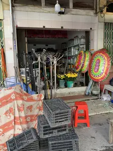 Đây là hình ảnh phía trước một cửa hàng bán đồ gia dụng ở Việt Nam. Cửa hàng đang mở cửa và trưng bày nhiều loại hàng hóa ra vỉa hè để bán. Các vật thể chính trong ảnh bao gồm: nhiều cây treo quần áo bằng kim loại (cả loại inox và sơn đen), hai vòng hoa lớn hình bầu dục với nhiều màu sắc rực rỡ được bọc trong nilon, và các xô nhựa đựng hoa cúc vàng. Ở phía trước có một chồng sọt nhựa màu đen và một chiếc ghế nhựa màu đỏ. Bên trong cửa hàng, có thể thấy các kệ sắt chứa đầy những chiếc bình thủy tinh lớn. Trên một cây cột ở bên trái có dán giấy ghi hai số điện thoại: 0974 331 565 và 0983 133 394.。（日本語訳）。これはベトナムにある家庭用品店の正面の写真です。店は営業中で、販売のために多くの種類の商品を歩道に陳列しています。写真の中の主な物体には、多くの金属製の衣類ハンガーラック（ステンレス製と黒色塗装の両方）、ナイロンで包まれた鮮やかな多色の大きな楕円形の花輪が2つ、そして黄色い菊の花が入ったプラスチック製のバケツが含まれます。手前には、黒いプラスチック製のカゴの山と、赤いプラスチック製の椅子が1脚あります。店内には、大きなガラス製の瓶でいっぱいの鉄製の棚が見えます。左側の柱には、2つの電話番号が書かれた紙が貼られています：0974 331 565と0983 133 394。