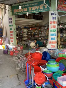 Bức ảnh chụp một cửa hàng kinh doanh tổng hợp, vừa là tiệm ảnh viện vừa bán đồ gia dụng, tại một khu vực ở Việt Nam. Cửa hàng có mặt tiền mở, bày bán hàng hóa tràn ra cả vỉa hè.。**Chữ viết được ghi lại:**。*   Biển hiệu chính màu xanh lá cây: "ẢNH VIỆN ÁO CƯỚI HOÀN TUYẾT" và các dịch vụ "CHỤP ẢNH NGHỆ THUẬT, ẢNH CƯỚI, ẢNH SINH NHẬT, ẢNH THẺ, HỘ CHIẾU, LỄ HỘI...".。*   Biển quảng cáo dọc trên cột bên trái: "CHỤP ẢNH SỰ KIỆN, PHÔNG RẠP, LOA ĐÀI, CƯỚI HỎI TRỌN GÓI".。*   Biển quảng cáo dọc trên cột bên phải: "PHỤC CHẾ ẢNH CŨ, CHỤP ẢNH THẺ LẤY NGAY, PHOTOCOPY, BÁN HỒ SƠ XIN VIỆC, LÀM KHUNG ẢNH CÁC LOẠI".。*   Biển hiệu màu đỏ ở góc trên bên phải (thấy một phần): "...SỰ KIỆN... QUAY CAMERA, PHỤC CHẾ ẢNH CŨ - ĐC: THÔN THẮNG TÂY - XÃ NGƯ...".。**Mô tả cảnh vật và hoạt động:**。*   **Loại địa điểm:** Cửa hàng tạp hóa kết hợp studio ảnh (ảnh viện).。*   **Hoạt động:** Một người phụ nữ đang ngồi xổm bên trong cửa hàng để sắp xếp hàng hóa. Bà đang cầm một chiếc nón lá.。*   **Các vật thể trong ảnh:** Rất nhiều đồ gia dụng bằng nhựa và kim loại được bày bán, bao gồm xô, chậu, rổ, kệ inox, thùng giữ nhiệt, và dụng cụ dọn dẹp. Bên trong, các kệ hàng chứa đầy bát đĩa, ấm chén, và phích nước. Không gian cửa hàng có nhiều hàng hóa được xếp chồng lên nhau.。（日本語訳）。ベトナムのある地域にある、写真館と家庭用品店を兼ねた総合商店を写した写真です。店は間口が広く、商品は歩道にまで溢れるように陳列されています。**記載されている文字：**。*   緑色のメイン看板：「HOAN TUYET ウェディングドレス写真館」およびサービス内容「芸術写真、結婚写真、誕生日写真、証明写真、パスポート、祭り…」。*   左の柱にある縦長の広告看板：「イベント撮影、背景幕、スピーカー、結婚式一式」。*   右の柱にある縦長の広告看板：「古い写真の修復、証明写真即時撮影、コピー、履歴書販売、各種額縁製作」。**風景と活動の説明：**。*   **場所の種類：** 雑貨店兼写真スタジオ（写真館）。*   **活動：** 店内で女性が一人、商品を整理するためにしゃがんでいます。彼女はノンラー（菅笠）を手に持っています。*   **写真の中の物：** プラスチック製や金属製の家庭用品が多数販売されており、バケツ、たらい、かご、ステンレス棚、クーラーボックス、掃除用具などが含まれます。店内では、棚に食器、茶器、魔法瓶がぎっしりと並んでいます。店のスペースには多くの商品が積み重ねられています。