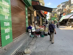 Bức ảnh chụp cảnh một con phố nhỏ ở Việt Nam, nơi diễn ra một phiên chợ cóc vào buổi sáng. Dọc theo con đường là những ngôi nhà ống san sát nhau. Bên trái, một cửa hàng đang đóng cửa cuốn màu nâu, bên cạnh có một tấm biển lớn màu xanh lá cây với dòng chữ 'DÙNG THUỐC AN TOÀN HIỆU QUẢ'. Trên vỉa hè, một người phụ nữ đang sắp xếp hàng hóa để bán, bao gồm hoa quả như bưởi và các nông sản khác được bày trên bàn và trong các rổ nhựa. Một người đàn ông lớn tuổi đang đi bộ trên đường, đeo túi chéo và tay cầm máy ảnh. Phía xa hơn trên con phố, có thể thấy các mái hiên của các cửa hàng khác, một người đang đi xe máy, và một lá cờ đỏ sao vàng của Việt Nam đang bay. Các biển hiệu khác có thể đọc được bao gồm 'TAXI GIA ĐÌNH' với tên 'NGUYỄN TRUNG' và các số điện thoại '0976 598 479', '0376 846 366', và một biển hiệu 'QUẦY THUỐC TRẦN NHÀN'.。（日本語訳）。ベトナムの小さな通りの風景を写した写真で、そこでは朝に路上市場が開かれています。道沿いには、チューブハウスが隣接して建ち並んでいます。左側には、茶色のシャッターが閉まっている店があり、その隣には「安全で効果的な薬の使用」という文字が書かれた大きな緑色の看板があります。歩道では、一人の女性が商品を並べて販売しており、それにはポメロなどの果物やその他の農産物がテーブルの上やプラスチックの籠の中に陳列されています。年配の男性が道を歩いており、ショルダーバッグを斜めがけにし、手にはカメラを持っています。通りのさらに奥には、他の店の軒先、バイクに乗っている人、そしてはためいているベトナムの金星紅旗が見えます。他に読み取れる看板には、「NGUYỄN TRUNG」という名前と電話番号「0976 598 479」、「0376 846 366」が書かれた「ファミリータクシー」、そして「TRẦN NHÀN薬局」という看板があります。