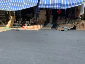 Hình ảnh chụp phía trước một cửa hàng có vẻ là cửa hàng tạp hóa hoặc bán đồ gia dụng, nằm ven một con đường nhựa. Cửa hàng được che bởi một mái hiên bạt lớn có sọc xanh và trắng, trên đó có thể đọc được chữ "BẠT TỰ CUỐN". Hàng hóa được bày la liệt từ trong ra ngoài thềm. Có thể nhận thấy nhiều thùng carton, một số ghi rõ "CUCKOO ELECTRIC RICE COOKER", cho thấy cửa hàng bán nồi cơm điện. Ngoài ra còn có rất nhiều nồi niêu, xoong chảo bằng kim loại xếp chồng lên nhau, các loại bình, phích giữ nhiệt. Bên trong cửa hàng, có một tấm biển nhỏ ghi "SỬA ĐỒNG HỒ". Phía bên phải là một đống vỏ thùng carton và một chiếc xô sơn cũ. Khung cảnh cho thấy một hoạt động kinh doanh buôn bán các mặt hàng đa dạng.。（日本語訳）。舗装された道路沿いにある、雑貨店または家庭用品店と思われる店の正面を撮影した写真。店は青と白の縞模様の大きな防水シートの日よけで覆われており、その上には「BẠT TỰ CUỐN」という文字が読み取れる。商品は店内から店先の土間までずらりと並べられている。多くの段ボール箱が見受けられ、その一部には「CUCKOO ELECTRIC RICE COOKER」と明記されており、この店が炊飯器を販売していることがわかる。その他にも、積み重ねられた金属製の鍋やフライパン、様々な種類の水筒や魔法瓶が多数ある。店内には、「SỬA ĐỒNG HỒ」と書かれた小さな看板がある。右側には、空の段ボール箱の山と古いペンキのバケツがある。この光景は、多様な商品を商う事業活動を示している。