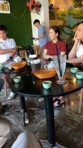 Bối cảnh là một không gian trong nhà, có thể là một quán trà hoặc quán cà phê. Một nhóm người đang ngồi tụ tập, nói chuyện quanh một chiếc bàn tròn màu đen. Trên bàn có các ấm trà sứ trắng, nhiều chén trà nhỏ màu xanh ngọc, một cốc thủy tinh cao và các giỏ đựng đồ. Mọi người dường như đang uống trà và ăn hạt hướng dương, vì có vỏ hạt vương vãi trên bàn và dưới sàn nhà. Trong số những người có mặt, một người phụ nữ mặc áo polo đỏ đang mỉm cười, một người đàn ông mặc áo polo trắng, và một người đàn ông khác mặc áo sơ mi xanh đang đứng ở phía sau. Trên tường phía sau họ có treo một chiếc quạt điện màu cam, một bức tranh phong cảnh lớn, và một tấm biển hiệu màu vàng chỉ đọc được một phần chữ 'HỒ'.。（日本語訳）。背景は屋内の空間で、茶館かカフェかもしれません。一団の人々が黒い円卓を囲んで座り、話をしています。テーブルの上には、白い磁器の急須、翡翠色の小さな茶碗が多数、背の高いガラスのコップが一つ、そして物入れ用の籠があります。皆はお茶を飲み、ひまわりの種を食べているようです。なぜなら、テーブルの上と床に種の殻が散らばっているからです。その場にいる人々の中には、赤いポロシャツを着て微笑んでいる女性が一人、白いポロシャツを着た男性が一人、そしてその後ろには青いシャツを着た別の男性が立っています。彼らの後ろの壁には、オレンジ色の扇風機、大きな風景画、そして「HỒ」という文字の一部しか読めない黄色い看板が掛かっています。