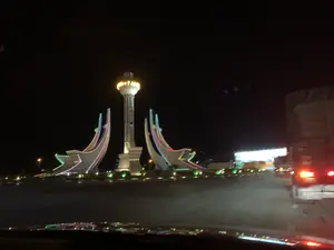 Bức ảnh chụp cảnh quan vào ban đêm, nhìn từ bên trong một chiếc xe ô tô đang di chuyển. Tại trung tâm của một vòng xuyến hoặc ngã tư lớn là một công trình kiến trúc điêu khắc hiện đại, được chiếu sáng rực rỡ. Công trình này bao gồm một tòa tháp cao ở giữa, có phần đỉnh phát sáng màu vàng, trông giống như một ngọn hải đăng. Hai bên tòa tháp là hai cấu trúc lớn, màu trắng, có hình dáng cách điệu của mũi thuyền (ghe/tàu) đang vươn ra. Các cấu trúc hình thuyền này được trang trí bằng các dải đèn LED nhiều màu sắc. Xung quanh công trình là đường nhựa và cây cảnh. Phía bên phải của bức ảnh có thể thấy phần đuôi của một chiếc xe tải lớn đang bật đèn. Phía sau công trình có các tòa nhà và ánh đèn khác nhưng bị mờ. Không có chữ viết nào có thể đọc được. Khung cảnh cho thấy đây là một công trình biểu tượng tại một nút giao thông quan trọng, có thể là cổng chào hoặc quảng trường của một địa phương ven biển, tôn vinh ngành hàng hải hoặc đánh bắt cá.。（日本語訳）。夜景を撮影した写真で、移動中の車内から見たものである。ロータリーまたは大きな交差点の中心には、明るくライトアップされた現代的な彫刻建築物がある。塔の両側には、船（小舟/船舶）の船首を様式化した形をした、前方に突き出ている大きな白い構造物が2つある。これらの船形の構造物は、色とりどりのLEDライトストリップで装飾されている。建造物の周りには、アスファルトの道路と観賞用の植物がある。建造物の背後には他の建物や灯りがあるが、ぼやけている。読み取れる文字はない。この光景は、これが重要な交通の要所にある象徴的な建造物であり、おそらくは沿岸地域の歓迎ゲートまたは広場で、海運業や漁業を称えるものであることを示している。