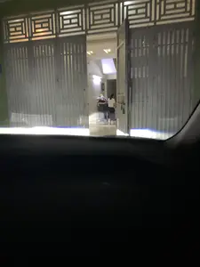 Ảnh chụp từ bên trong một chiếc ô tô vào ban đêm. Đèn pha của xe đang chiếu sáng vào một ngôi nhà có cổng sắt lớn màu xám. Cánh cổng có hoa văn hình học ở phía trên và đang mở một phần, để lộ không gian sáng đèn bên trong. Một người mặc áo trắng đang đứng trong nhà, quay lưng lại. Cảnh vật cho thấy một ngôi nhà dân vào buổi tối.。（日本語訳）。夜、車の中から撮影された写真。車のヘッドライトが、大きな灰色の鉄の門がある家を照らしている。門の上部には幾何学模様があり、一部が開いていて、中の明るく照らされた空間が見えている。白い服を着た人が家の中に立っており、背中を向けている。この光景は、夜の民家を示している。