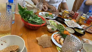 Cảnh một bữa ăn trong nhà, có nhiều người đang ngồi quây quần bên một chiếc bàn gỗ. Trên bàn bày biện thịnh soạn nhiều món ăn Việt Nam, bao gồm một đĩa tôm luộc, một đĩa thịt luộc (hoặc lòng), một đĩa lạc rang, một đĩa sung muối, một rổ rau sống và bánh đa nướng. Có nhiều loại nước chấm được đặt trong các bát nhỏ. Về đồ uống, có các lon bia Hà Nội, lon Coca-Cola và một chai nước khoáng hiệu "THỦY SƠN". Mọi người đang cùng nhau ăn uống, tạo nên một không khí sum họp, gần gũi.。（日本語訳）。家での食事の光景、多くの人々が木製のテーブルを囲んで座っている。テーブルの上には、茹でエビ一皿、茹で肉（またはモツ）一皿、煎りピーナッツ一皿、イチジクの塩漬け一皿、生野菜一かご、そして焼きライスペーパーなど、多くのベトナム料理が豪華に並べられている。飲み物については、ハノイビールの缶、コカ・コーラの缶、そして「THỦY SƠN」ブランドのミネラルウォーターのボトルがある。皆が一緒に飲食しており、団らんの親密な雰囲気を醸し出している。