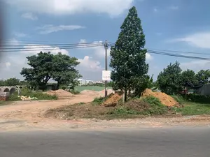 Trên một cột điện có một tấm biển quảng cáo của một cửa hàng vật liệu xây dựng, ghi các mặt hàng như: 'CÁT - ĐÁ - GẠCH - XI MĂNG', 'ĐẤT MÀU - ĐẤT PHÙ SA'. Tên cửa hàng ở trên cùng không đọc được rõ. Khung cảnh là một bãi đất trống ven đường, được sử dụng làm nơi tập kết vật liệu xây dựng. Có nhiều đống cát và đất, cùng với các ống cống bê tông xếp chồng lên nhau. Cây cối, cột điện và dây điện cũng xuất hiện trong ảnh. Ở phía xa, sau một hàng rào công trình màu xanh, là một tòa nhà cao tầng màu trắng. Bầu trời trong xanh, có mây. Con đường nhựa nằm ở phía trước.。（日本語訳）。電柱の上に、ある建材店の広告看板があり、「砂 - 石 - レンガ - セメント」、「色のついた土 - 沖積土」といった品目が書かれている。一番上にある店名は、はっきりと読めない。風景は道端の空き地で、建材の集積場所として使われている。砂と土の山がたくさんあり、コンクリートの土管が積み重ねられている。木々、電柱、そして電線も写真に写っている。遠くには、青い工事用のフェンスの後ろに、白い高層ビルがある。空は青く澄んでおり、雲がある。手前にはアスファルトの道がある。