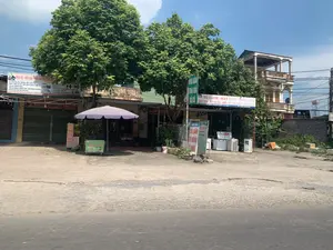 Khung cảnh một dãy cửa hàng ven đường vào một ngày nắng. Có nhiều cây xanh lớn tỏa bóng mát. Bên trái là một cửa hàng có biển hiệu "MUA HỘ - ĐẤU GIÁ VÀN CHUYÊN HÀNG N...", chuyên kinh doanh các mặt hàng như "Túi Xách Hàng Hiệu: MICHAEL KORS, GUCCI, COACH, LOUIS VUITTON...", "Giày Dép: ADIDAS, NIKE...", "Quần Áo: UNIQLO, ADIDAS, ZARA...", và "Hàng Điện Tử: Apple: IPHONE, WATCH, AIRPODS...". Ở giữa là một cửa hàng điện máy có biển hiệu lớn màu xanh và trắng ghi: "TỦ LẠNH - MÁY GIẶT - ĐIỀU HÒA. BÁN TRAO ĐỔI ĐỒ CŨ - MỚI - ĐỒ GIA DỤNG - BÀN - GHẾ". Địa chỉ được ghi là "Đ/C: XÃ THẠCH HÒA - THẠCH THẤT - HÀ NỘI" và số điện thoại "ĐT: 0355 202 082". Phía trước cửa hàng trưng bày nhiều tủ lạnh, máy giặt và điều hòa đã qua sử dụng. Ngoài ra còn có các biển quảng cáo nhỏ ghi "MUA BÁN TRAO ĐỔI ĐỒ CŨ" và "TỦ LẠNH, TỦ ĐÔNG, TỦ MÁT". Phía trước một cửa hàng khác có một chiếc ô lớn màu tím. Gần đó có một xe đẩy bán nước "MÍA" và một tấm biển nhỏ ghi "CHAN GA" (chân gà), "BÁNH TRÁNG". Phía sau là một ngôi nhà nhiều tầng với ban công. Con đường phía trước là đường nhựa.。（日本語訳）。晴れた日の道端に並ぶ店の風景。大きな緑の木々がたくさんあり、日陰を作っている。左側には「MUA HỘ - ĐẤU GIÁ VÀN CHUYÊN HÀNG N...」（代理購入 - オークション、N...商品専門）という看板を掲げた店があり、「ブランドバッグ：マイケル・コース、グッチ、コーチ、ルイ・ヴィトン...」、「靴：アディダス、ナイキ...」、「衣類：ユニクロ、アディダス、ザラ...」、そして「電子機器：アップル：iPhone、Watch、AirPods...」などの商品を専門に扱っている。中央には、青と白の大きな看板を掲げた電器店があり、「冷蔵庫 - 洗濯機 - エアコン。中古品 - 新品 - 家電 - テーブル - 椅子 の販売・交換」と書かれている。住所は「Đ/C: XÃ THẠCH HÒA - THẠCH THẤT - HÀ NỘI」（住所：ハノイ市タックタット県タックホア社）、電話番号は「ĐT: 0355 202 082」（電話：0355 202 082）と記されている。店の前には、多くの中古の冷蔵庫、洗濯機、エアコンが陳列されている。その他にも、「中古品の売買・交換」や「冷蔵庫、冷凍庫、冷蔵ショーケース」と書かれた小さな広告看板がある。別の店の前には、大きな紫色のパラソルがある。その近くには、「MÍA」（サトウキビジュース）を売る屋台と、「CHAN GA」（鶏の足）、「BÁNH TRÁNG」（ライスペーパー）と書かれた小さな看板がある。後ろにはバルコニー付きの多層階の家がある。前の道はアスファルトの道路である。