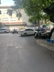 Trên một tấm bạt màu xanh treo trên cây có dòng chữ: 'LÊ ĐIỆN', 'CÁC LOẠI Ô, VÕNG - CHIẾU' và số điện thoại '☎ 0918 109 75...'.。Bối cảnh là một con đường hoặc một con hẻm trong một khu đô thị. Dọc theo con đường trải nhựa là các phương tiện đang đỗ, bao gồm cả ô tô và xe máy. Một số xe máy đậu trên vỉa hè được phủ bạt che. Có một người đang điều khiển xe tay ga di chuyển trên đường. Ở phía bên phải, dưới một gốc cây lớn, có hai chiếc ghế nhựa nhỏ màu xanh được đặt trên vỉa hè, có thể là nơi để ngồi nghỉ hoặc một quán nước nhỏ. Phía sau là một tòa nhà nhiều tầng màu vàng.。（日本語訳）。木に掛けられた青いシートの上には、「LÊ ĐIỆN」、「各種傘、ハンモック - マット」、そして電話番号「☎ 0918 109 75...」という文字があります。背景は、ある都市部の一本の道または路地です。舗装された道に沿って、自動車やバイクを含む車両が駐車しています。歩道に停められたいくつかのバイクは、カバーシートで覆われています。スクーターを運転している人が一人、道を移動しています。右側には、大きな木の根元の下、歩道に2つの小さな青いプラスチックの椅子が置かれており、休憩する場所か、あるいは小さな飲み物屋かもしれません。後ろには、黄色い多層階の建物があります。