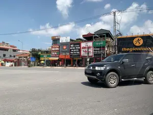 Bức ảnh chụp một ngã tư đường phố sầm uất ở Sơn Tây vào ban ngày, dưới trời nắng đẹp. Một chiếc ô tô SUV màu đen đang chạy qua, hình ảnh hơi mờ do chuyển động. Dọc hai bên đường là những tòa nhà phố nhiều tầng, được sử dụng làm cửa hàng kinh doanh.。Các hoạt động chính là giao thông và buôn bán. Có một vài người đang đi xe máy và nhiều xe máy khác đang đậu trước các cửa hàng.。Các biển hiệu quảng cáo và thông tin có thể đọc được bao gồm:。- Chuỗi cửa hàng lớn: 'thegioididong.com' với biển hiệu lớn màu vàng đen.。- Các thương hiệu điện thoại: 'MI STORE' (Xiaomi), 'OPPO'.。- Các cửa hàng địa phương: 'SIÊU THỊ ĐIỆN THOẠI THỊNH CƯỜNG II', 'ĐIỆN THOẠI THỊNH CƯỜNG', 'KÍNH MẮT VIỆT AN', 'PHONE SHOP'.。- Dịch vụ: 'Bán Trả Góp', 'ĐIỆN THOẠI - LAPTOP - LINH PHỤ KIỆN', 'SỬA CHỮA - THAY MÀN - ÉP KÍNH', 'CHUYỂN TIỀN NHANH'.。- Địa danh: Một biển hiệu ghi địa chỉ tại 'SƠN LỘC - SƠN TÂY - HÀ NỘI'. Cổng một ngôi trường có tên 'TRƯỜNG ... SƠN LỘC'.。- Biển báo giao thông: Biển báo màu xanh 'CHÚ Ý' quy định hướng đi cho các loại xe ô tô.。Ngoài ra, trong ảnh còn có hệ thống cột điện, dây điện chằng chịt, đèn tín hiệu giao thông và vạch kẻ đường.。（日本語訳）。写真は、晴天の昼下がり、ソンタイ（Sơn Tây）の賑やかな交差点を撮影したものです。一台の黒いSUV車が通り過ぎており、その動きによって画像は少しぼやけています。道路の両脇には、商業店舗として利用されている多層階のタウンハウスが並んでいます。主な活動は交通と商業です。数人がバイクに乗っており、他の多くのバイクが店の前に停められています。読み取れる広告や情報の看板には、以下のものが含まれます：。- 大手チェーン店：「thegioididong.com」、黄色と黒の大きな看板があります。- 携帯電話ブランド：「MI STORE」（Xiaomi）、「OPPO」。- 地元の店：「SIÊU THỊ ĐIỆN THOẠI THỊNH CƯỜNG II」、「ĐIỆN THOẠI THỊNH CƯỜNG」、「KÍNH MẮT VIỆT AN」、「PHONE SHOP」。- サービス：「Bán Trả Góp」（分割払い販売）、「ĐIỆN THOẠI - LAPTOP - LINH PHỤ KIỆN」（携帯電話 - ノートパソコン - アクセサリー）、「SỬA CHỮA - THAY MÀN - ÉP KÍNH」（修理 - 画面交換 - ガラス圧着）、「CHUYỂN TIỀN NHANH」（スピード送金）。- 地名：「SƠN LỘC - SƠN TÂY - HÀ NỘI」という住所が書かれた看板。「TRƯỜNG ... SƠN LỘC」という名前の学校の門。- 交通標識：「CHÚ Ý」（注意）と書かれた青い標識が、各種自動車の進行方向を規定しています。