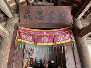 Bối cảnh là một gian thờ cúng trong một công trình kiến trúc cổ của Việt Nam, có thể là đình làng hoặc nhà thờ họ, với kết cấu chủ yếu bằng gỗ như cột, kèo và mái nhà lợp ngói. Phía trên cùng của gian thờ có một bức hoành phi bằng gỗ, trên đó khắc nổi bốn chữ Hán: 緩家思成 (Hoãn Gia Tư Thành). Hai bên cột của gian thờ có treo hai câu đối bằng gỗ. Câu đối bên phải (nhìn từ ngoài vào) có các chữ 庭左右 (Đình Tả Hữu). Câu đối bên trái có các chữ 減春秋 (Giảm Xuân Thu). Phía trước bàn thờ có treo một tấm yếm lụa màu đỏ, được thêu hình rồng và hoa lá tinh xảo, có các tua rua nhiều màu sắc. Trên bàn thờ bày các đồ thờ cúng như lọ hoa cắm hoa giả, một vòng nhang lớn hình xoắn ốc, chân nến, và các vật phẩm khác. Không gian thể hiện hoạt động tín ngưỡng thờ cúng tổ tiên hoặc thần linh.。（日本語訳）。背景はベトナムの古い建築物の中にある祭壇で、村の亭（ディン）か一族の祠堂（ nhà thờ họ）である可能性がある。主な構造は柱、梁、瓦葺きの屋根など木材でできている。祭壇の最上部には木製の扁額（ホアン・フィ）があり、そこには「緩家思成（ホアン・ザー・トゥ・タイン）」という四つの漢字が浮き彫りにされている。祭壇の両側の柱には、木製の二つの対聯（ついれん）が掛けられている。（外から見て）右側の対聯には「庭左右（ディン・ター・ヒュウ）」という文字がある。左側の対聯には「減春秋（ザム・スアン・トゥ）」という文字がある。祭壇の前には赤い絹の幕（イエム）が掛けられており、精巧な龍と花葉の刺繍が施され、色とりどりの房飾りが付いている。この空間は、祖先または神々を祀る信仰活動を表している。
