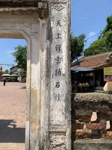 Bức ảnh chụp một góc nhìn từ trong ra ngoài của một cổng đá cổ, có thể là cổng làng Mông Phụ tại làng cổ Đường Lâm, Sơn Tây. Bối cảnh là một ngày nắng đẹp.。**Chữ viết:**。1.  Trên cột cổng bên phải, có một hàng chữ Hán được khắc dọc màu đen: 天上祥雲銷石栓 (Thiên thượng tường vân tiêu thạch thuyên).。2.  Ở phía bên phải, có một tấm biển quảng cáo màu vàng của cửa hàng tên 'Hương Sen' với nội dung: "CHO THUÊ VÀ BÁN SẴN QUẦN ÁO DÀI TRANG PHỤC, ÂU PHỤC Truyền Thống Làng Cổ Đường Lâm. ĐC: ĐÌNH MÔNG PHỤ - XÃ ĐƯỜNG LÂM - SƠN TÂY. ĐT: 0985 940 310".。**Bối cảnh và hoạt động:**。*   **Địa điểm:** Một di tích lịch sử ở làng cổ Đường Lâm, Sơn Tây. Cảnh vật bao gồm một cổng đá lớn, cũ kỹ với các chi tiết chạm khắc; một bức tường xây bằng đá ong; và một khoảng sân rộng lát gạch đỏ.。*   **Hoạt động:** Bên ngoài cổng, có một cửa hàng nhỏ cho thuê trang phục truyền thống (áo dài) và các loại trang phục khác cho du khách chụp ảnh. Một vài người đang ngồi nghỉ dưới mái che của cửa hàng. Một người phụ nữ đang đi bộ trên sân gạch.。*   **Vật thể:** Cột cổng bằng đá đã phong hóa, tường đá ong, sân gạch đỏ, mái nhà lợp ngói âm dương, biển quảng cáo, bàn ghế, và vài chiếc nón lá đặt trên bàn.。（日本語訳）。写真は、古い石の門の内側から外側を向いた視点を捉えたもので、ソンタイのドゥオンラム古村にあるモンフー村の門である可能性があります。背景は美しい晴れの日です。**文字：**。1.  右側の門柱には、「天上祥雲銷石栓」（Thiên thượng tường vân tiêu thạch thuyên）という漢詩が黒色で縦に刻まれています。2.  右側には、「Hương Sen」という店の黄色い広告看板があり、内容は次の通りです。「ドゥオンラム古村の伝統的なアオザイ衣装、洋服のレンタルおよび既製品販売。住所：ソンタイ、ドゥオンラム社、モンフー亭。電話：0985 940 310」。**背景と活動：**。*   **場所：** ソンタイのドゥオンラム古村にある歴史的遺跡。風景には、彫刻が施された大きくて古い石の門、ラテライトで造られた壁、そして赤レンガで舗装された広い中庭が含まれています。*   **活動：** 門の外には、観光客が写真を撮るために伝統衣装（アオザイ）やその他の衣装を貸し出す小さな店があります。数人が店の軒下で座って休んでいます。一人の女性がレンガ敷きの中庭を歩いています。*   **物体：** 風化した石の門柱、ラテライトの壁、赤レンガの中庭、陰陽瓦で葺かれた屋根、広告看板、テーブルと椅子、そしてテーブルの上に置かれた数個のノンラー（ベトナムの円錐形の帽子）。