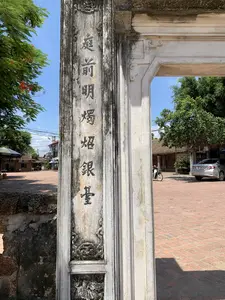 Bức ảnh chụp cận cảnh một bên cột của một chiếc cổng đá cổ, có lẽ là cổng của một đình làng hoặc một di tích lịch sử ở Sơn Tây. Trên thân cột có khắc một hàng chữ Hán theo chiều dọc: 庭前明燭銀臺 (phiên âm Hán-Việt: Đình tiền minh chúc ngân đài). Cột đá trông cũ kỹ, nhuốm màu thời gian, với các chi tiết hoa văn chạm khắc tinh xảo nhưng đã bị bào mòn. Nhìn qua khung cổng là một khoảng sân rộng được lát bằng gạch đỏ. Trong sân, một chiếc ô tô màu bạc đang đỗ và có một người đang đi xe máy. Phía xa là những ngôi nhà kiến trúc cũ với mái ngói. Bên trái bức ảnh là tán lá xanh của cây phượng với những chùm hoa đỏ, nổi bật trên nền trời xanh trong. Khung cảnh tổng thể mang vẻ yên bình, tĩnh lặng của một không gian văn hóa vào một ngày nắng đẹp.。（日本語訳）。古い石造りの門の片側の柱のクローズアップ写真。おそらくソンタイにある村の亭（てい）、または歴史的遺跡の門であろう。柱の本体には、縦書きで一行の漢文が刻まれている。「庭前明燭銀臺」（漢越語読み：Đình tiền minh chúc ngân đài）。石柱は古びており、時の流れを感じさせる色合いで、精巧に彫られた模様の細部は摩耗している。門の枠を通して見ると、赤レンガが敷き詰められた広い中庭がある。庭には銀色の車が一台駐車しており、バイクに乗っている人が一人いる。写真の左側には、赤い花房をつけたホウオウボク（鳳凰木）の緑の葉冠があり、澄んだ青空を背景に際立っている。全体的な光景は、晴れた美しい日における文化空間の平穏で静かな趣を帯びている。