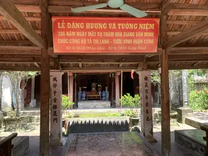 Bức ảnh chụp bên trong một khu di tích lịch sử có kiến trúc cổ, dường như là một đền thờ hoặc nhà thờ họ. Một tấm băng rôn lớn màu đỏ với chữ vàng được treo trang trọng trên xà nhà bằng gỗ, ghi nội dung: "LỄ DÂNG HƯƠNG VÀ TƯỞNG NIỆM 386 NĂM NGÀY MẤT CỤ THÁM HOA GIANG VĂN MINH, CHỨC CÔNG BỘ TẢ THỊ LANG - TƯỚC VINH QUẬN CÔNG, 02/6/1638 (Mậu Dần) - 02/6/2024 (Giáp Thìn)". Hai bên lối vào gian thờ chính là hai cột trụ lớn có khắc câu đối bằng chữ Hán: cột bên trái ghi "風聲千古探花門" (Phong thanh thiên cổ Thám hoa môn) và cột bên phải ghi "禮義百年蒙阜邑" (Lễ nghĩa bách niên mông phụ ấp). Nhìn sâu vào bên trong là ban thờ được bài trí trang nghiêm với lư hương, hoa cúng và các bình gốm sứ trang trí (lộc bình). Phía trước ban thờ có một bể nước nhỏ. Toàn bộ khung cảnh cho thấy sự chuẩn bị hoặc diễn ra của một buổi lễ tưởng niệm. Các chi tiết khác bao gồm mái ngói, cột kèo gỗ, quạt trần và nền gạch hoa văn cổ.。（日本語訳）。古代建築様式の史跡内部で撮影された写真で、寺院または一族の祠堂のようです。木造の梁に、金色の文字が書かれた大きな赤い横断幕が厳かに掲げられており、その内容は「タムホア・ザン・ヴァン・ミン公、職：工部左侍郎、爵：栄郡公の没後386周年（1638年6月2日（戊寅）- 2024年6月2日（甲辰））献香・追悼式典」と記されています。本堂への入り口の両側には、漢文の対句が刻まれた2本の大きな柱があります。左の柱には「風聲千古探花門」（風声千古探花門）、右の柱には「禮義百年蒙阜邑」（礼義百年蒙阜邑）と記されています。奥に目をやると、香炉、供花、装飾用の陶磁器の壺（福禄瓶）で厳かに飾られた祭壇があります。祭壇の前には小さな水盤があります。全体の光景は、追悼式典の準備または開催中であることを示しています。その他の細部には、瓦屋根、木製の柱と梁、天井扇風機、そして古い模様のタイル張りの床が含まれます。