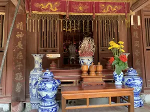 Bức ảnh chụp một không gian thờ cúng trang nghiêm, có khả năng là tiền sảnh của một ngôi đình, đền hoặc chùa với kiến trúc bằng gỗ. Ở trung tâm là một ban thờ được bài trí công phu. Phía trước có một bàn gỗ, trên đó đặt một lọ hoa sứ men lam trắng cắm hoa cúc vàng và hoa mẫu đơn đỏ. Xung quanh ban thờ là các bình gốm sứ trang trí lớn (lộc bình) men lam trắng với hoa văn phong cảnh. Trên ban thờ chính, đặt trên một bệ gỗ cao hơn, có một lư hương lớn bằng sứ, bên trong cắm rất nhiều que hương đang cháy. Bên cạnh lư hương là các vật phẩm thờ cúng khác như ba hũ gỗ nhỏ có nắp và một đĩa lễ đặt trên đôn, trên đó có một hộp bánh quy. Phía trên lối vào gian thờ chính có một tấm rèm vải màu đỏ (y môn) được trang trí với viền và tua rua màu vàng, trên đó thêu hình rồng vàng và chữ Hán. Hai bên lối vào là hai cột gỗ lớn có khắc các câu đối bằng chữ Hán màu vàng. Chữ trên câu đối có thể đọc được một phần: cột bên trái là "精神長存子孫..." (Tinh thần trường tồn tử tôn...) và cột bên phải là "敬蹟同垂南邦..." (Kính tích đồng thùy Nam bang...). Khung cảnh cho thấy một hoạt động thờ cúng, thể hiện qua việc hương đang cháy và các lễ vật được dâng lên.。（日本語訳）。写真は、木造建築の村の集会所、祠、または寺の前堂と思われる荘厳な祭祀空間を写している。中央には、精巧に飾られた祭壇がある。祭壇の周りには、風景模様が描かれた青と白の染付の大きな装飾用の陶磁器の壺（福禄瓶）がある。主祭壇の上、より高い木製の台の上には、大きな磁器の香炉があり、その中には燃えている線香がたくさん挿してある。香炉の隣には、蓋付きの小さな木製の壺3つや、高坏に置かれた供物皿（その上にはクッキーの箱が一つある）といった他の祭具がある。主祭壇への入り口の上には、黄色い縁取りと房で装飾された赤い布の垂れ幕（y môn）があり、その上には金色の龍と漢字が刺繍されている。入り口の両側には、金色の漢字で対聯が刻まれた2本の大きな木の柱がある。対聯の文字は一部読むことができる。左の柱は「精神長存子孫...」（Tinh thần trường tồn tử tôn...）、右の柱は「敬蹟同垂南邦...」（Kính tích đồng thùy Nam bang...）である。この光景は、燃えている線香や捧げられた供物を通して表される祭祀活動を示している。